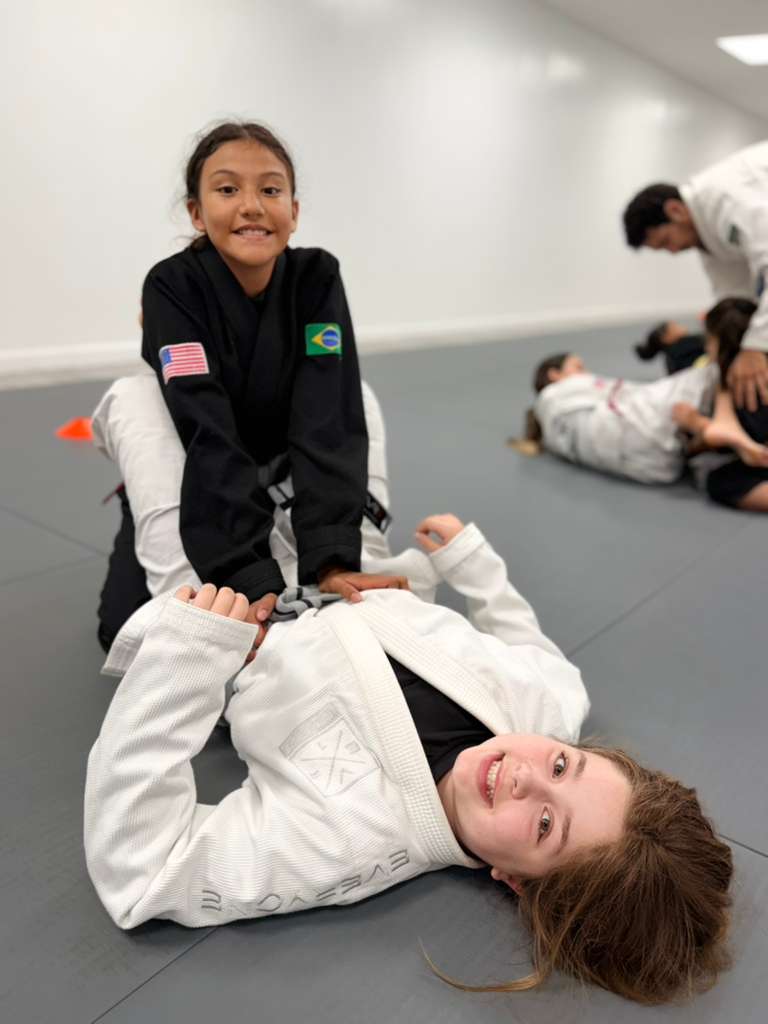 Two young students smiling on the mats while drilling jiu-jitsu together during class