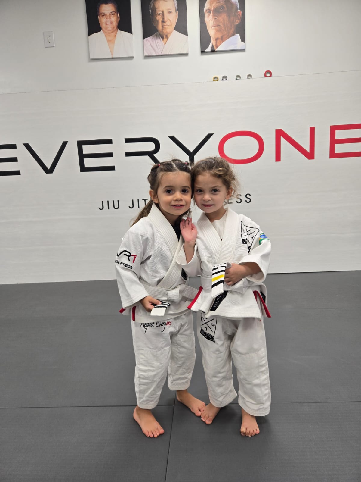 Two young students in white gis smiling in front of the Everyone Jiu Jitsu wall logo