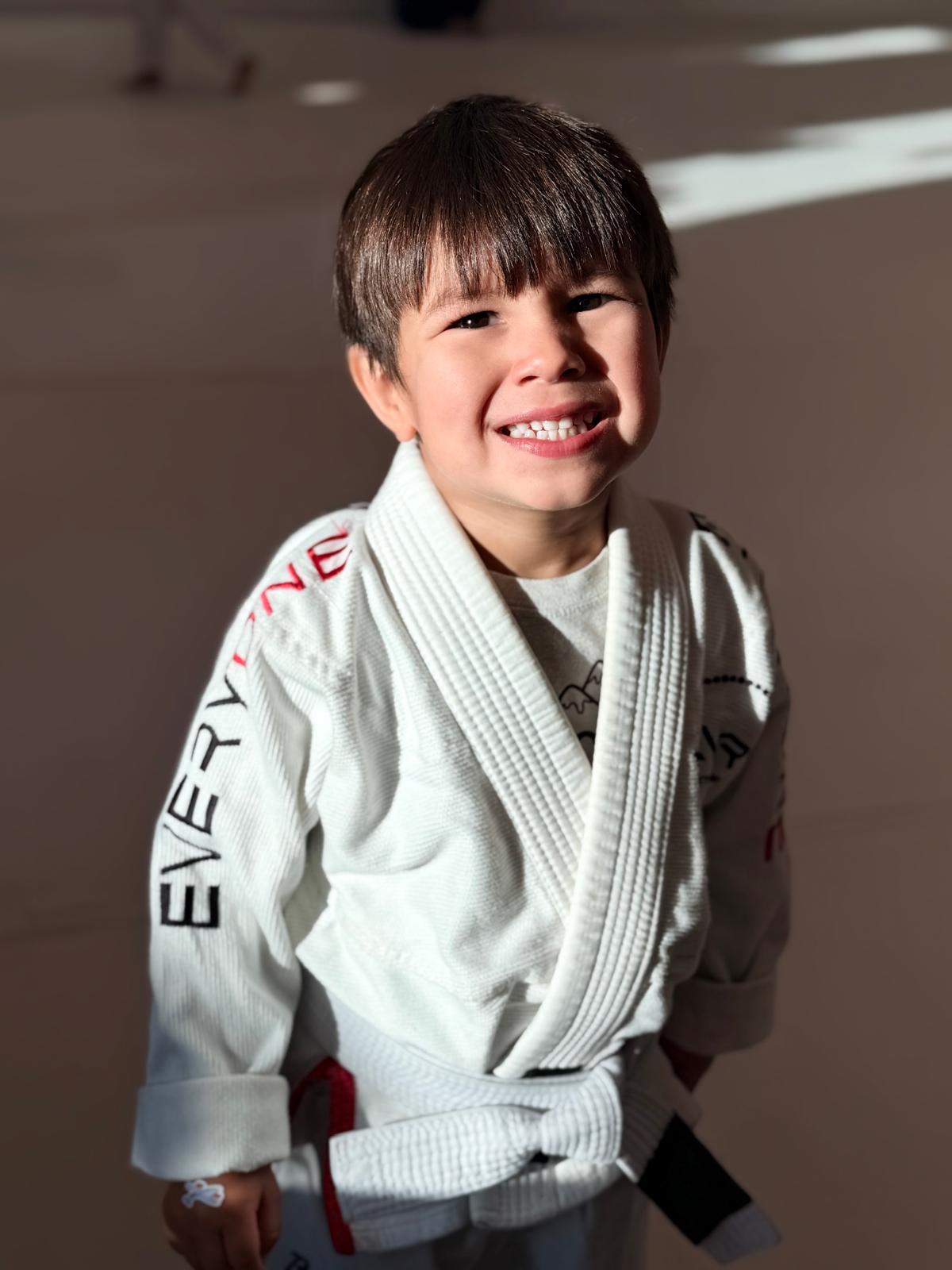 A young student in a white Everyone Jiu Jitsu gi smiling on the academy mats